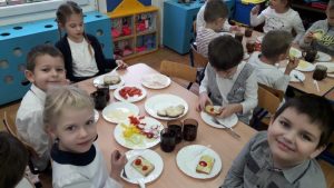 Kilkoro dzieci siedzi na krześle przy stole. Na stole leżą talerze, pokrojone warzywa, chleb z masłem i kubek z herbatą.