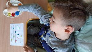 Dziecko siedzi przy stoliku. Na kartce papieru maluje palcami kolorowe kropki.