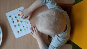 Dziecko siedzi przy stoliku. Na kartce papieru maluje palcami kolorowe kropki.