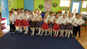 Dzieci stoją w 2 rzędach. Z przodu dziewczynki ubrane w spódniczki w kwiaty, z tyłu chłopcy w białych koszulach