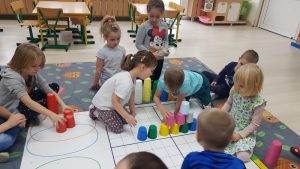 Kilkoro dzieci siedzących na dywanie. W środku znajduje się mata, na której ułożona jest wieża z kolorowych kubków. Dwoje dzieci trzyma w ręku plastikowe kubki. Jeden chłopiec układa z kubków wieżę.