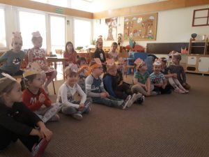 Grupa dzieci siedzących w półkolu na dywanie. Za nimi troje dzieci i Pani siedzący na krzesłach.