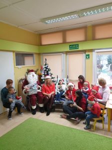 Grupa dzieci z nauczyelkami siedzi na krześle lub wózku. Na środku siedzi Mikołaj. Za Mikołajem stoi choinka.