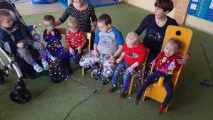 Grupa dzieci i 2 Panie siedzą na krzesłach. w rękach trzymaja kolorowej lampki choinkowe.