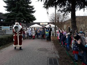 Grupy dzieci stojących w ogrodzie przedszkolnym. Przed nimi stoi Mikołaj przemawiający przez megafon