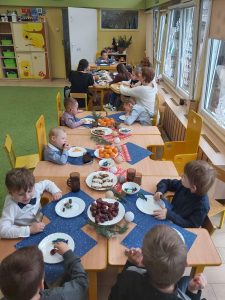 Grupa dzieci siedząca przy stole. Na stołach znajdują się talerze, pierniki, ciastka, mandarynki, serwetki.