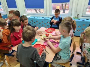 Kilkoro dzieci siedzi przy stoliku. Przed sobą mają tacę z wyciętym szablonem piernikowego ludzika. Trzymają w ręce pędzelek, a niektóre posypkę po pierników.