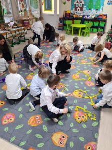 Grupa dzieci siedzących na dywanie wraz z trzema Paniami. Dzieci układają kształty z kolorowych pasków bibuły.
