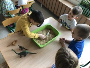 Czworo chłopców siedzących przy stolikach. Na stoliku znajduje się pojemnik z piaskiem.Jeden chłopiec patrzy przez lupę i trzyma rękę w pojemniku z piaskiem.