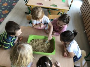 Dzieci siedzące przy stoliku. Po środku stołu znajduje się pojemnik z piaskiem. Dziewczynka trzyma w ręku pędzel umieszczony w piasku.