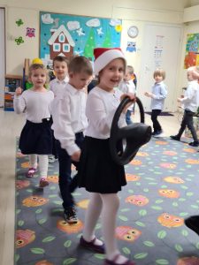 Dzieci na dywanie ustawione w kole jedno za drugim. Na pierwszym planie dziewczynka trzymająca kierownicę.