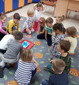 Dzieci siedzące w kole na dywanie. Po środku koła znajduję się taca z pojemnikiem z niebieską pianą.
