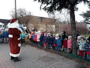 Grupa dzieci stojących w ogrodzie przedszkolnym wraz z dwoma Paniami. Przed nimi stoi Mikołaj.
