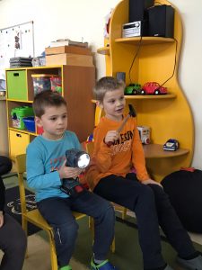 Dwóch chłopców siedzących na krzesłach. Chłopiec w niebieskiej bluzce trzyma w ręku latarkę, natomiast chłopiec w pomarańczowej bluzce trzyma w ręku młotek.