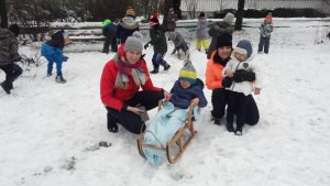 Dziecko siedzi na sankach, które trzyma Pani . Obok stoi dziewczynka trzymana przez Panią. Za nimi grupa dzieci rzuca się snieżkami. Na dworze jest dużo śniegu.