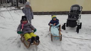 Dwoje dzieci siedzi na sankach. Za nimi stoi dziewczynka ze śnieżką w ręce.