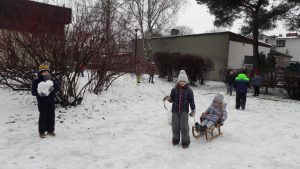 Dziewczynka trzyma sznurek od sanek, na których siedzi inna dziewczynka. Obok stoi chłopiec z dużą kulką śniegu.