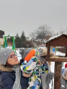 Dziecko jest trzymane na rękach przez Panią. Sięga ręką do karmnika. Sypie do niego ziarna.