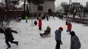Grupa dzieci na placu przedszkolnym. Jedne stoją, inne pochylają się po śnieg. Na środku siedzi dziecko na sankach. Jest dużo śniegu.