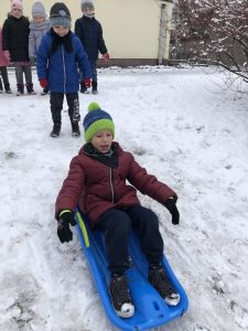 Chłopiec zjeżdżający na sankach w przedszkolnym ogrodzie. Za nim stoi czworo dzieci.