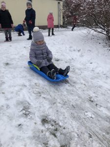 Dziewczynka zjeżdżająca na sankach w przedszkolnym ogrodzie. Za nią stoi troje dzieci. Jedno dziecko schyla się po śnieg.