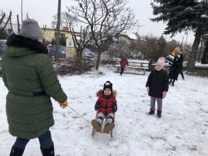 Pani ciągnąca dziecko na sankach w zimowym ogrodzie. Za nimi stoi dziewczynka i jeszcze jedna Pani. Z tyłu biegnie chłopiec.
