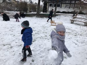 Dzieci i Panie bawiące się w pokrytym śniegiem ogrodzie przedszkolnym. Dziewczynka znajdująca się na pierwszym planie trzyma w ręku kulkę śniegu.