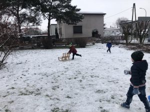 Troje dzieci bawiących się na śniegu w ogrodzie przedszkolnym. Jeden chłopiec popycha sanki.
