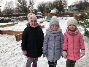 Trzy dziewczynki stojące obok siebie na pokrytym śniegiem przedszkolnym placu zabaw.