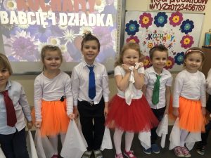 Sześcioro dzieci odświętnie ubranych, stojących w sali przedszkolnej w jednym rzędzie i trzymających w rękach białe, materiałowe chustki.