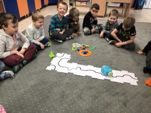 Siedmioro dzieci siedzących w półkolu na dywanie w sali przedszkolnej. Przed dziećmi znajduje się robocik poruszający sie po trasie ułożonej z biało-czarnych puzzli, plastikowe koło, wieża oraz kolorowe kostki.