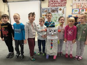 Ośmioro dzieci stojących w rzędzie w sali przedszkolnej. Przed dziećmi stoi robot wykonany z materiałów plastycznych.
