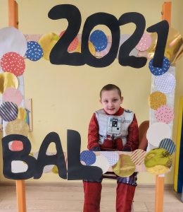 Chłopiec w przebraniu robota siedzi na krześle za dużą ramką ozdobioną koilorowymi kołami i napisem BAL 2021