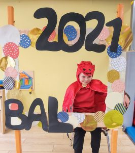 Chłopiec w przebraniu ( peleryna i czapka czerwona) siedzi na krześle za dużą ramką ozdobioną koilorowymi kołami i napisem BAL 2021
