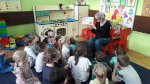 Grupa dzieci siedzi na dywanie. Na środku na krześle siedzi nauczycielka. Trzyma w rękach otwartą książkę.