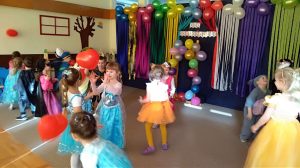 Dzieci tańczące w parach w sali przedszkolnej i odbijające balony. Dzieci mają ubrane karnawałowe stroje. W tle, na ścianie znajduje się dekoracja złożona z balonów i kolorowych pasków bibuły.