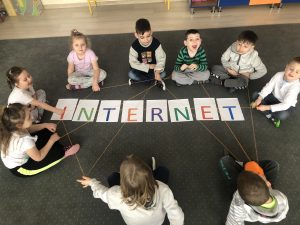 Grupa dzieci siedzących w kole na dywanie i trzymających w rękach wełnę tworzącą sieć. W środku, na podłodze znajduje się napis INTERNET.