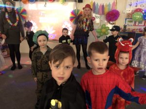 Na pierwszym planie znajduje się czterech chłopców ubranych w karnawałowe kostiumy. Na drugim planie znadują się dwie panie w kapeluszach oraz czworo dzieci ubranych w karnawałowe stroje.