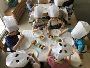 Sześcioro dzieci ubranych w fartuszki i czapki kucharskie siedzący przy stolikach, na których znajdują się talerze i owoce. Dzieci wykonują owocowe szaszłyki.