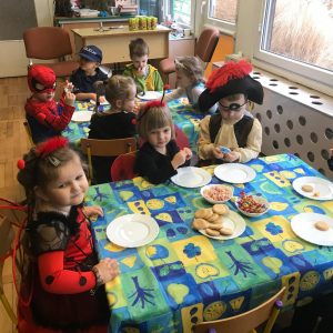 Dzieci siedzące przy stolikach w sali przedszkolnej. Przedszkolaki ubrane są w karnawałowe stroje, na stołach znajdują się talerze oraz ciastka i cukierki.