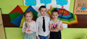 3 dzieci ( jeden chłopiec po środku i dwie dziewczynki) stoją. Dziewczynki trzymają w ręce kolorowe parasole