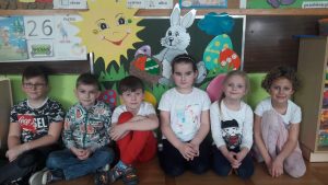 6 dzieci siedzi na ziemi. Za nimi na tablicy umieszczony jest papierowy króliczek, koszyczek, pisanki, słońce.