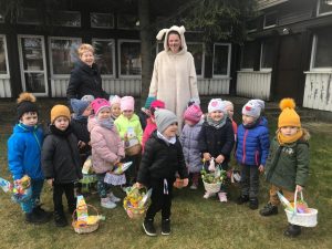 Grpa dzieci pozująca do zdjęcia w ogrodzie przedszkolnym i trzymająca w rękach wielkanocne koszyczki. Za dziećmi stoją dwie Panie. Pani po prawej stronie ma założony strój zajączka