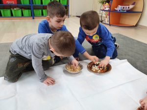 Troje dzieci siedzących na dywanie w sali przedszkolnej i biorących z talerzyków groch.