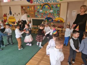 Grupa dzieci tańczących w parach w sali przedszkolnej wraz z dwoma Paniami. Dzieci mają na głowach ubrane kolorowe czapeczki.