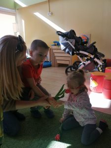 Chłopiec wraz z Panią wręczający siedzącej na dywanie dziewczynce tulipana.