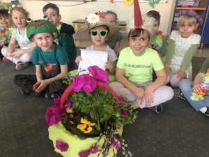 Dziewięcioro dzieci siedzących w sali przedszkolnej w dwóch rzędach.Dzieci w pierwszym rzędzie siedzą na dywanie. Dziewczynka siedząca w środku ma założone przeciwsłoneczne okulary i wiosenny kapelusz. Chłopiec siedzący po lewej stronie ma na głowie czapkę - żabkę. Chłopiec po prawej stronie ma na głowie czapkę - bociana. Chłopcy siedzacy w drugim rzędzie mają naszyjach naszyjniki z kwiatów, dziewczynki wiosenne opaski. Przed dziećmi stoi koszyk z ziołami i kwiatkami.