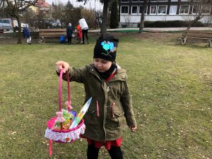 Na pierwszym planie dziewczynka trzyma w ręce wielkanocny koszyczek. Z tyłu widocznych jest czworo dzieci wraz z Pania szukających koszyczków wielkanocnych.