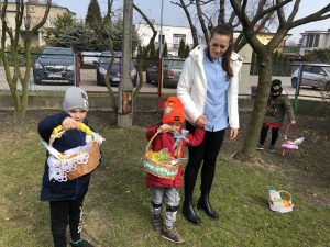 Na pierwszym planie, w ogrodzie przedszkolnym stoi dwoje dzieci i tzryma w rękach wielkanocne koszyczki. Chłopiec po prawej stronie trzyma za rękę Panią. Z tyłu za drzewem stoi dziewczynka trzymająca w ręku koszyczek wielkanocny.