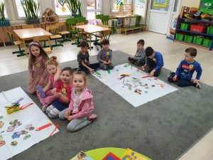 Dziewięcioro dzieci siedzących na dywanie w sali przedszkolnej. Przed dziećmi znajdują się materiały plastyczne służące do robienia papierowych zakładek do książek.